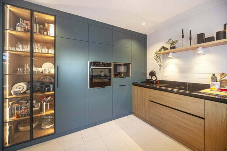 Blue-grey tall units with lit glass cabinet beside an oak kitchen run with black worktop